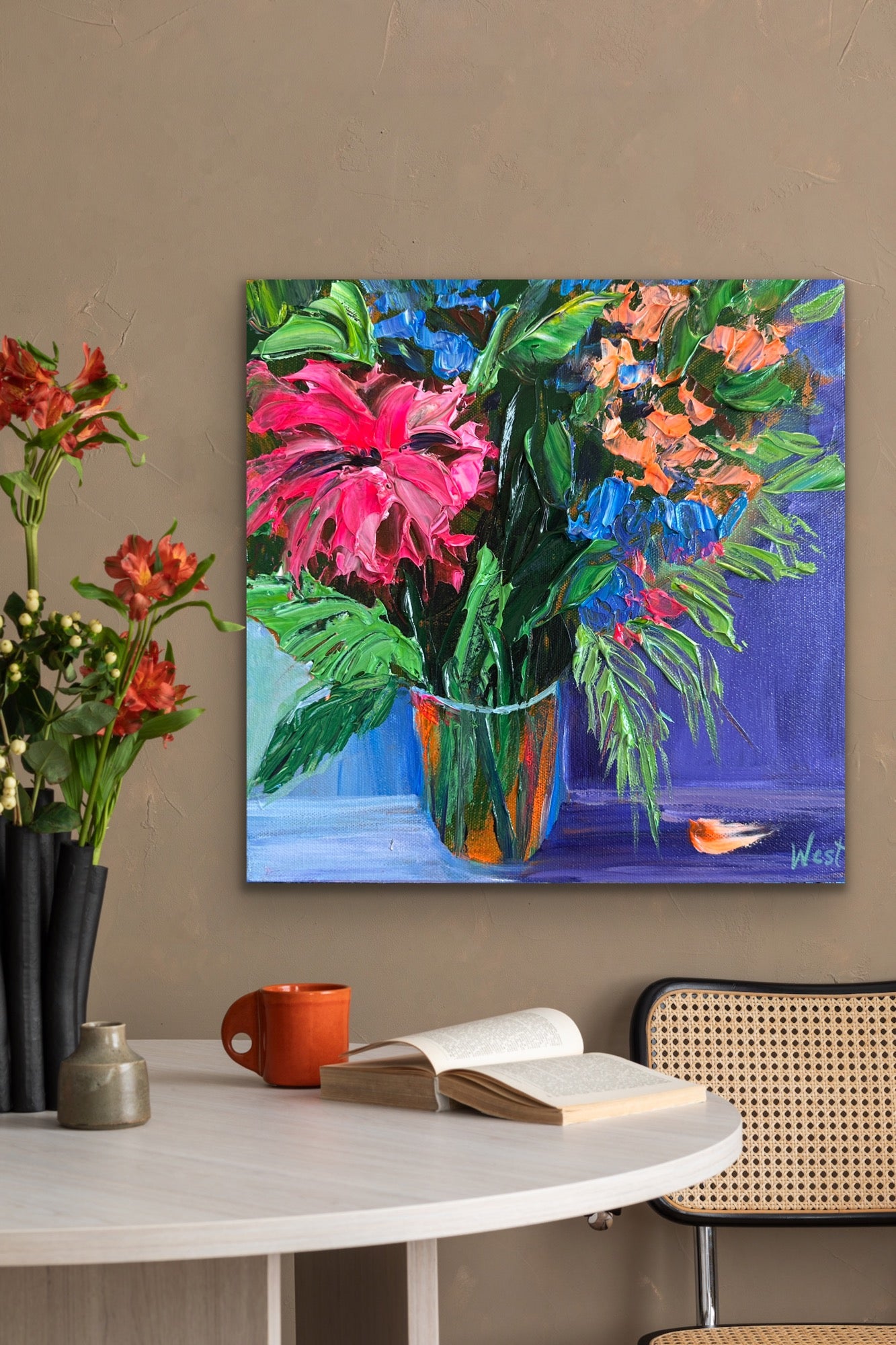 Colorful palette knife impasto painting of flowers on a wall above a table with a book and mug.