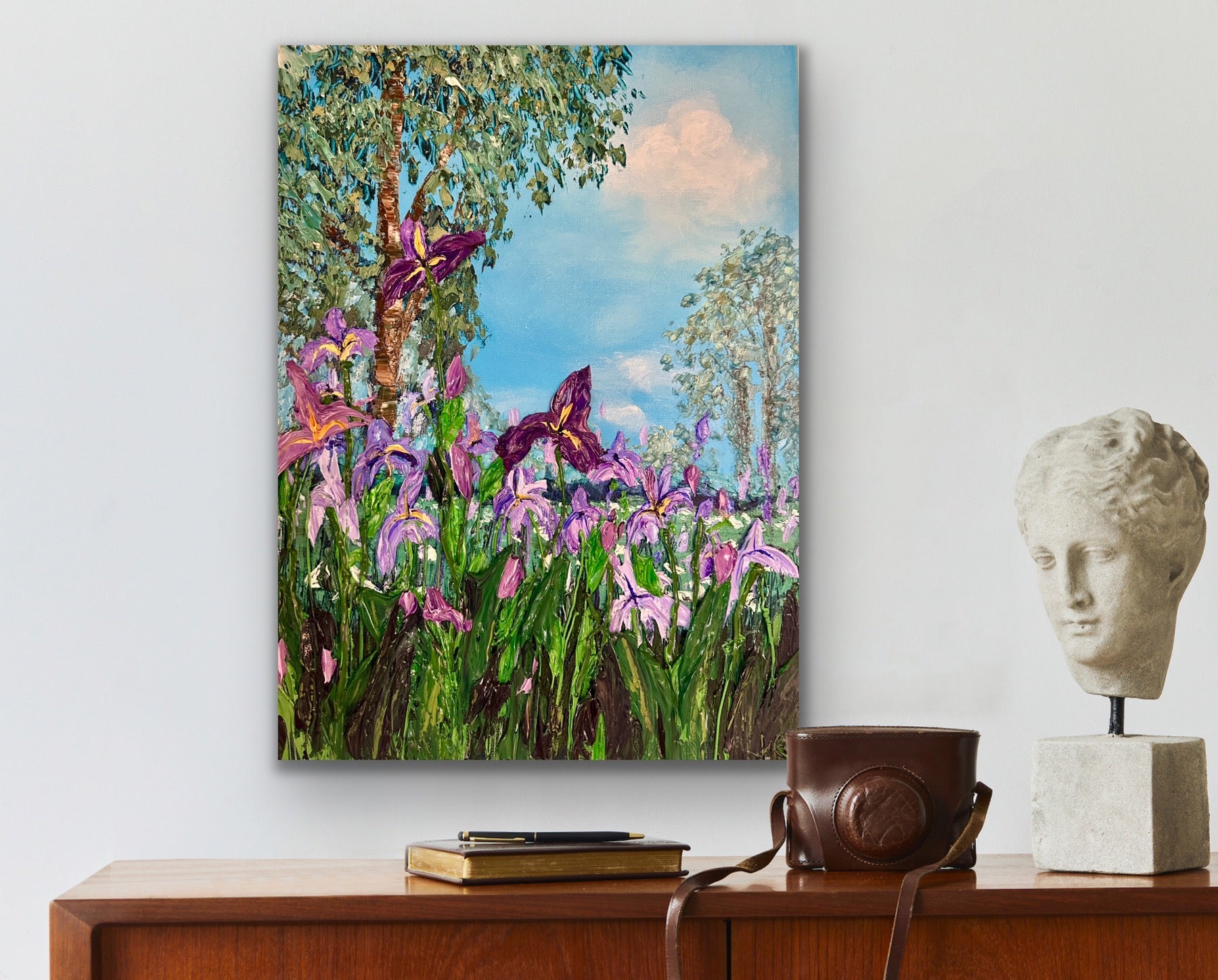 Painting of irises on a wall above a wooden cabinet with a camera and sculpture.