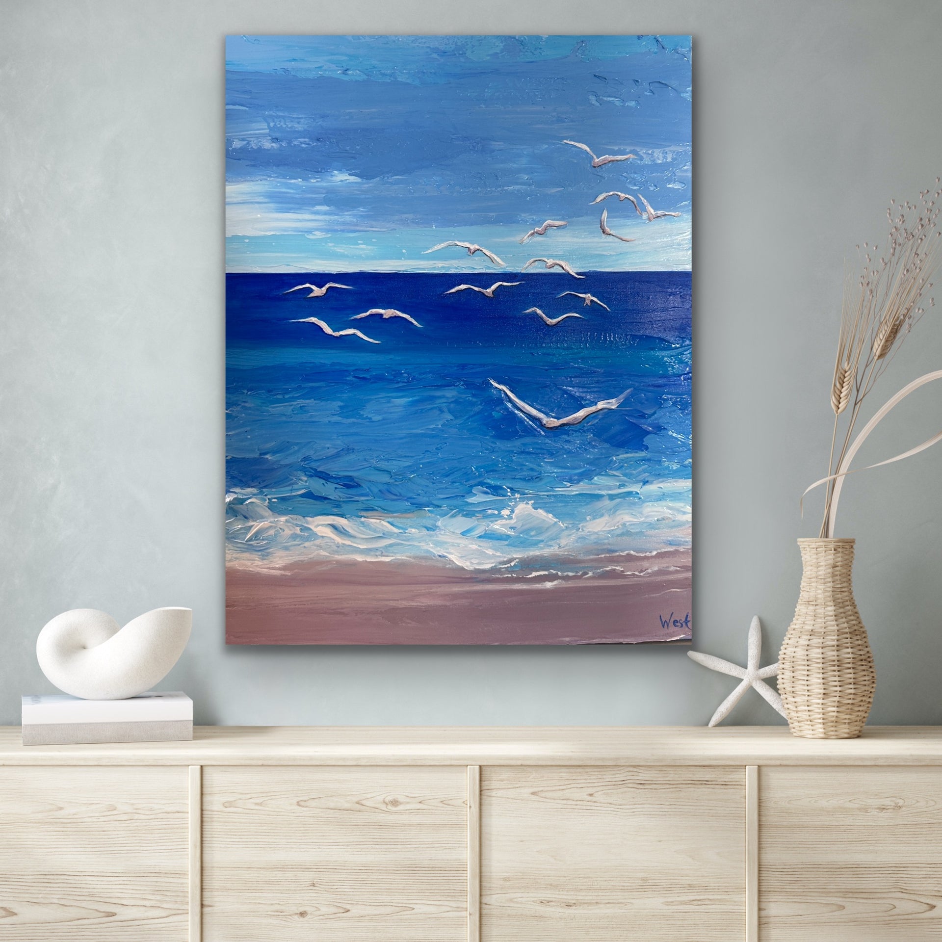 Palette knife impasto painting of a beach scene with blue sky, white clouds, and birds over water on a wall above a wooden shelf.